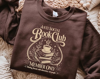 May include: A brown sweatshirt with the text "Anti Social Book Club" in a vintage style font. The design includes a stack of books, a coffee cup, and the words "Member Only: Making friends with characters, not people."