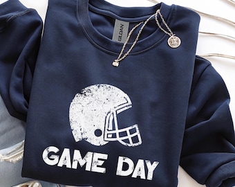 May include: Navy blue sweatshirt with a distressed white football helmet graphic and the words "GAME DAY." A silver necklace with a small pendant rests on the sweatshirt. The sweatshirt is folded neatly on a white surface.