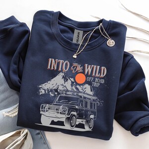 May include: Navy blue sweatshirt with a graphic design featuring a mountain range, a setting sun, and an off-road vehicle. The text "Into The Wild Off Road Trip" is printed above the vehicle. A silver necklace with a pendant is draped on the sweatshirt.