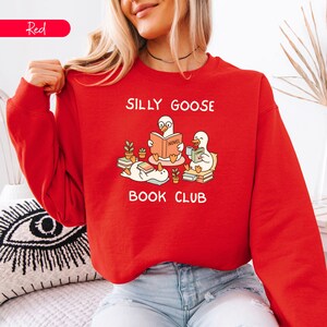 May include: A red sweatshirt featuring the words "SILLY GOOSE BOOK CLUB" and a graphic of three cartoon geese reading books. The geese are surrounded by small potted plants. The word "Red" is in a red rectangle in the upper left corner.