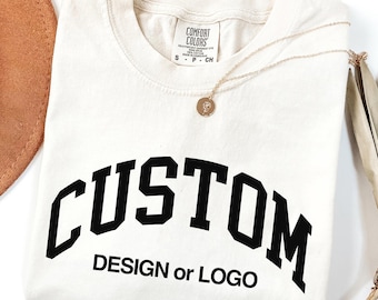 May include: A cream-colored short-sleeve t-shirt with a Comfort Colors brand label at the neck. The front features the black text CUSTOM in a large arched font, with the words DESIGN or LOGO printed in a smaller black sans-serif font below.