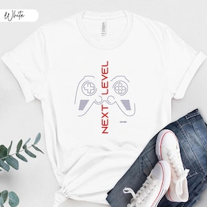 May include: White t-shirt with a blue and red graphic of a video game controller and the words "NEXT LEVEL" in red. The word "Gamer" is below the controller. The shirt is paired with blue jeans and white sneakers.