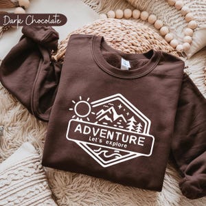 May include: Dark chocolate-colored sweatshirt with a white graphic that reads "ADVENTURE, Let's explore" with a sun, mountains, and trees design. The sweatshirt is folded on a woven surface with decorative beads.