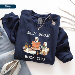 May include: Navy blue sweatshirt featuring the words "SILLY GOOSE BOOK CLUB" and a graphic of geese reading books. The sweatshirt has a crew neck and long sleeves. A silver necklace with a pendant is draped on the sweatshirt.