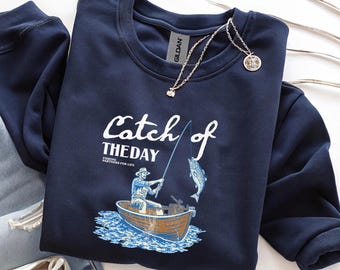 Peut inclure: Sweat-shirt bleu marine avec l'inscription "Catch of the Day" et un dessin d'un pêcheur dans une barque attrapant un poisson. Un collier argenté avec un pendentif est posé sur le sweat-shirt.