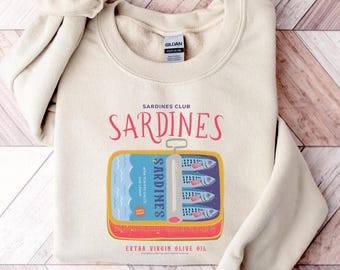 May include: A cream-coloured sweatshirt with a sardine can graphic. The design includes the words "Sardines Club" and "Sardines" in a whimsical font, plus "Extra Virgin Olive Oil". The can illustration shows sardines with tomato sauce and lemon.