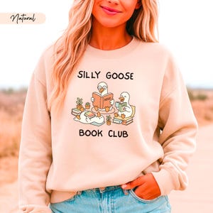 May include: A beige sweatshirt featuring the words "Silly Goose Book Club" and a graphic of cartoon geese reading books. The word "Natural" is in the upper left corner. The sweatshirt is a neutral colour.