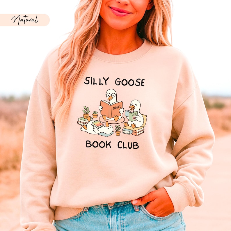 May include: A beige sweatshirt featuring the words "Silly Goose Book Club" and a graphic of cartoon geese reading books. The word "Natural" is in the upper left corner. The sweatshirt is a neutral colour.