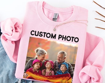 May include: A light pink crewneck sweatshirt featuring the black text CUSTOM PHOTO above a rectangular printed image of an adult and two children sitting together outdoors, with the children wearing colorful hats and wrapped in a bright blanket.