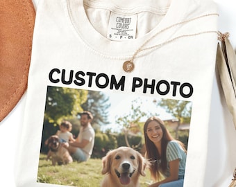 May include: A cream-colored short-sleeve t-shirt with a Comfort Colors label at the neck. The shirt features the black text CUSTOM PHOTO above a printed photograph showing a family with two dogs outdoors in a grassy area.