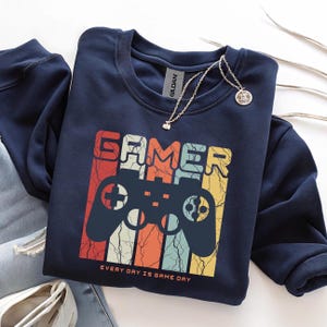 Könnte beinhalten: Marineblaues Sweatshirt mit einem Retro-Design mit der Aufschrift "GAMER" über einer Controller-Illustration. Der Text "EVERY DAY IS GAME DAY" steht darunter. Eine silberne Halskette mit Anhänger befindet sich auf dem Sweatshirt.