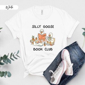 May include: White t-shirt featuring the text "SILLY GOOSE BOOK CLUB" and a cartoon illustration of geese reading books. The shirt is styled with blue jeans and white trainers.
