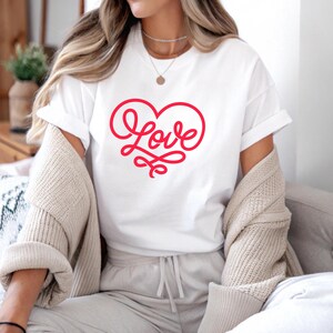 May include: White t-shirt with a red heart-shaped design that spells out the word "Love". The shirt has short sleeves and is made of a soft material. A beige sweater is draped over the shoulders.