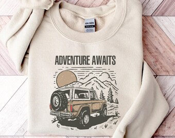 Puede incluir: Sudadera color crema con el texto "ADVENTURE AWAITS" sobre un gráfico de un vehículo todoterreno marrón, montañas y un sol. El diseño está impreso en tonos marrones y grises. La sudadera está doblada sobre una superficie de madera.