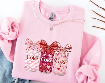 May include: A pale pink sweatshirt displaying a graphic of three Diet Coke cans decorated with bows. The cans feature various patterns, including hearts, stripes, and lips. A delicate necklace with a pendant is also visible.