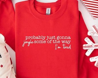 May include: A red sweatshirt with the text "probably just gonna jingle some of the way I'm tired" in white. The sweatshirt is folded and surrounded by holiday decorations, including a gift box and decorative snowflakes.