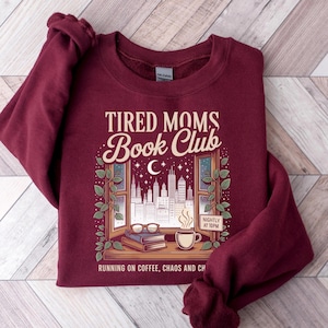Può includere: Una felpa bordeaux con la scritta "TIRED MOMS Book Club" sopra un'illustrazione di uno skyline cittadino visto attraverso una finestra. Il design include una luna crescente, stelle, libri, occhiali e una tazza di caffè. Il testo "RUNNING ON COFFEE, CHAOS AND CH" è in basso.