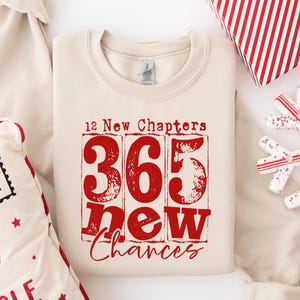 Könnte beinhalten: Cremefarbenes Sweatshirt mit rotem Text "12 New Chapters 365 New Chances". Der Text ist in einem rechteckigen Design angeordnet. Das Sweatshirt ist gefaltet, mit einer Geschenkbox und Dekorationen im Hintergrund.