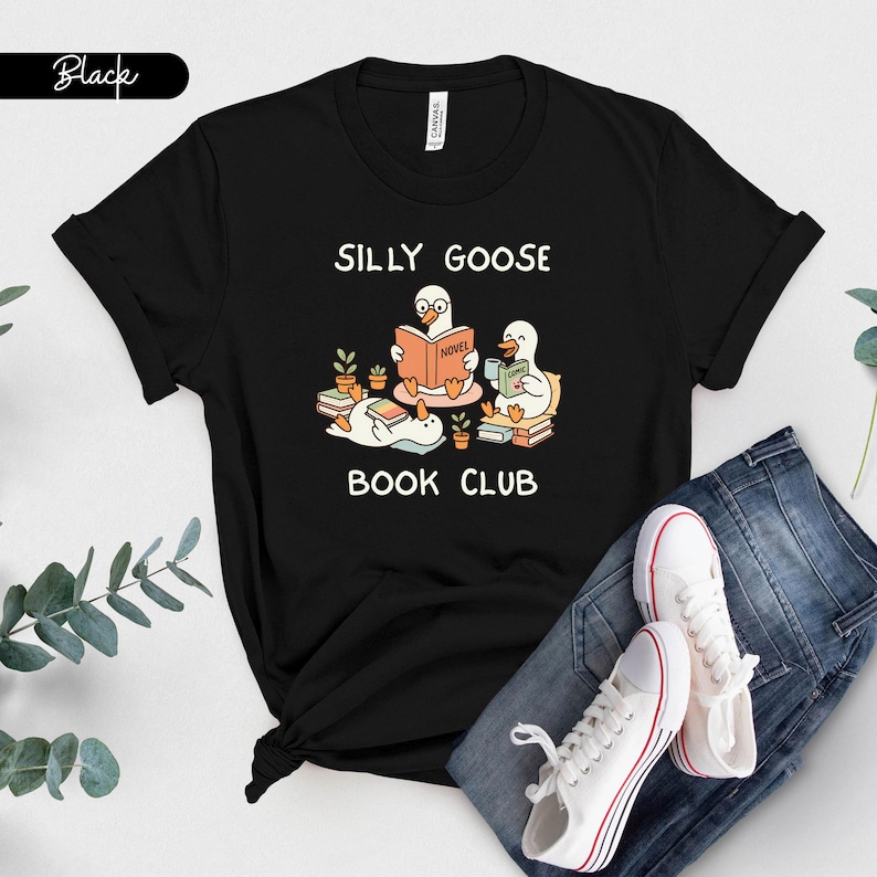 May include: Black t-shirt with a cartoon design of three geese reading books, with the words "SILLY GOOSE BOOK CLUB." The illustration includes potted plants and the word "NOVEL" on a book. White trainers and blue jeans are also visible.