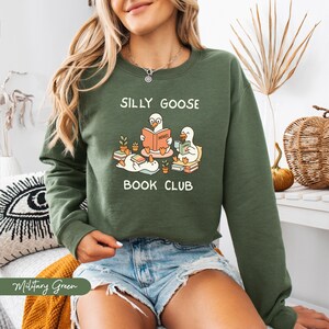 May include: A military green crew neck sweatshirt featuring the text "SILLY GOOSE BOOK CLUB" and a graphic of cartoon geese reading books. The sweatshirt is made of a soft material and is a casual style.