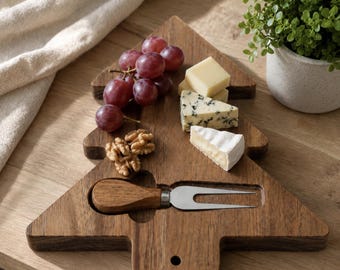 May include: A Christmas tree-shaped wooden serving board featuring a recessed slot holding a metal cheese fork with a wooden handle. The board is topped with red grapes, walnut halves, and three varieties of cheese, including blue cheese.