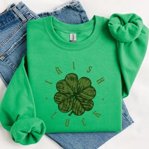 May include: A bright green sweatshirt with the words "Irish Luck" surrounding a four-leaf clover design. The sweatshirt is folded on top of a pair of blue jeans. The sleeves are scrunched up.
