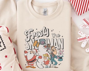 May include: Cream-colored sweatshirt featuring the text "Frosty the Snowman" with a graphic of Frosty, Santa, and other characters. The design is in a retro style with a vintage aesthetic. A festive holiday-themed apparel item.