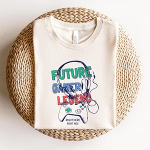 Può includere: T-shirt color crema con le parole "FUTURE GAMER LEGEND" in verde, blu e rosso, insieme a una grafica di un gamepad e cuffie. Il testo "RIGHT HERE RIGHT NOW" è sotto il gamepad. La maglietta è piegata su una superficie tessuta.