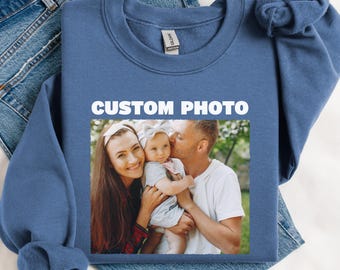 May include: A blue sweatshirt with the words "CUSTOM PHOTO" printed in white. The sweatshirt features a family photo of a woman, a man, and a baby. The sweatshirt is lying on a pair of blue jeans.