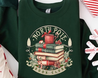 Puede incluir: Sudadera verde oscuro con un diseño gráfico que presenta una pila de libros, una taza roja y el texto "North Pole Book Club". El diseño incluye elementos festivos como bastones de caramelo y acebo, ideal para Navidad.
