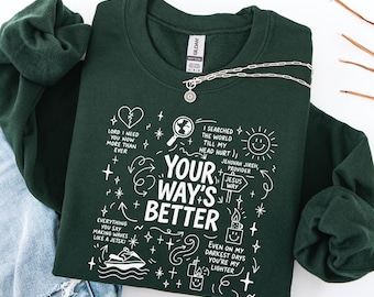 May include: A dark green sweatshirt with white graphic text that reads "Your Way's Better" and other phrases. The sweatshirt has a crew neck and long sleeves. A silver necklace with a pendant is draped across the sweatshirt.