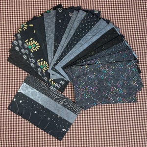May include: A collection of assorted fabric swatches in various shades of gray and black, arranged in a fan shape. The swatches feature different patterns, including floral, polka dots, and geometric designs. The background is a brown and white checkered pattern.