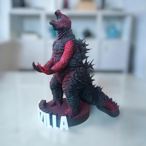 以下が含まれることがあります： ゴジラのようなモンスターの精巧なフィギュア。主に黒色で、胸、腕、頭に赤色のアクセントがあります。フィギュアはダイナミックなポーズで、ベースには「ZILLA」という名前が表示されています。コレクションアイテムを示唆するデザインです。