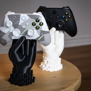 May include: Two 3D printed hands holding Xbox controllers. One controller is black and the other is white with a camouflage pattern and the text "POWER YOUR DREAMS" and "PROJECT XARLETTS".
