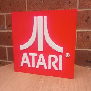 May include: A square, red sign displaying the Atari logo in white. The logo comprises three vertical lines and the word "ATARI" in a bold, sans-serif font. The registered trademark symbol is present.