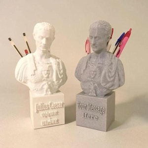 May include: Two bust-shaped desk organisers featuring Julius Caesar. The white bust displays the text "Julius Caesar 13 July 100 BC 15 March 44 BC" on its base. The grey bust displays the text "Your Message Here" on its base. Both hold pens.