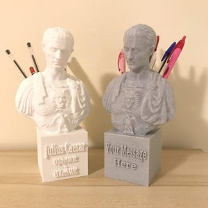 May include: Two pen holders shaped as Roman busts, one white and one grey. The white bust is labelled "Julius Caesar" with dates. The grey bust has the text "Your Message Here". Pens are placed in the busts. The busts are made of plastic.