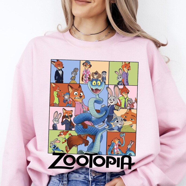 May include: A light pink sweatshirt featuring a colorful graphic of characters from the animated film "Zootopia." The design includes a grid of images with various characters and the word "Zootopia" in black block letters at the bottom.