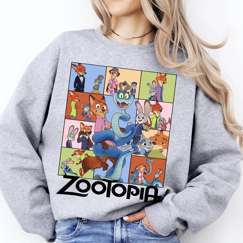 May include: A light gray sweatshirt featuring a colorful graphic from the animated film "Zootopia." The graphic is a grid of character illustrations, including a snake, fox, and bunny. The word "Zootopia" is printed in black at the bottom.