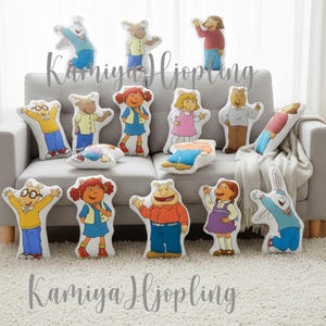 Puede incluir: Una colección de cojines con forma de personajes de la serie de televisión animada Arthur. Los cojines representan a los personajes en varias poses y atuendos, con colores brillantes y diseños detallados. Los cojines están colocados en un sofá gris.