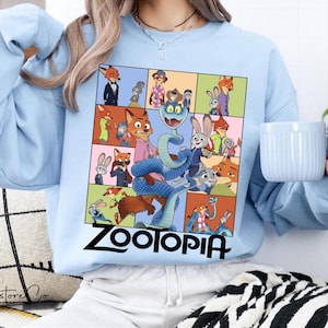 May include: Light blue sweatshirt featuring a colorful collage of characters from the animated film "Zootopia." The design includes images of Nick Wilde, Judy Hopps, and other characters, with the word "Zootopia" in bold black letters at the bottom.