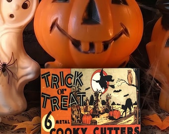 May include: A rectangular box labelled Trick or Treat 6 Metal Cooky Cutters featuring a graphic illustration of a witch, a black cat, and pumpkins. The box is positioned in front of two lit candles in brass holders, a white ghost figure, and two orange jack-o'-lanterns.