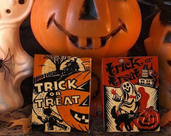 May include: A collection of Halloween decorations including a white ghost figure, two orange plastic jack-o'-lanterns, and two rectangular signs with the text Trick or Treat. Two brass candlesticks with lit candles are placed on either side.