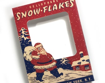 May include: A vintage red and white cardboard frame with a cutout for a photo. The frame features a winter scene with a Santa Claus carrying a bag of gifts, a house, and snow. The text "CELLOPHANE SNOW-FLAKES" is printed at the top of the frame. The text "STAR BAND CO. * NEW YORK, N.Y." is printed at the bottom of the frame.