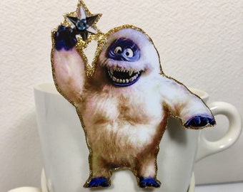 Puede incluir: Una taza de cerámica blanca con un borde de purpurina dorada y un recorte de papel del abominable hombre de las nieves sosteniendo una estrella. El abominable hombre de las nieves lleva botas azules y tiene un cuerpo blanco y peludo. La taza está encima de otra taza de cerámica blanca.