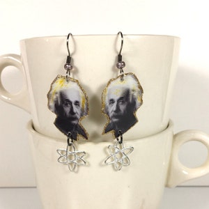 Puede incluir: Pendientes colgantes con una imagen en blanco y negro de Albert Einstein, un borde amarillo y dorado y abalorios de átomos plateados. Los pendientes cuelgan de ganchos plateados. Los pendientes se muestran frente a una taza blanca.