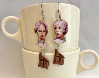 May include: A pair of dangle earrings featuring a chocolate bar charm and a printed image of a woman with a pink hat. The earrings are hanging from a white mug.