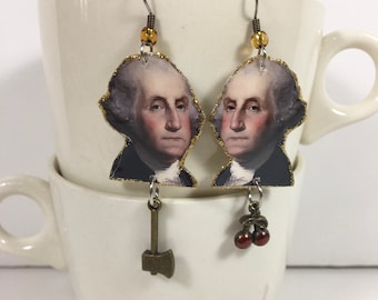 Puede incluir: Un par de pendientes con un retrato de George Washington en purpurina dorada. Un pendiente tiene un pequeño dije de hacha, el otro tiene dos dijes de cereza roja.