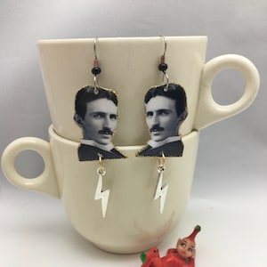 May include: A pair of silver earrings featuring a black and white image of Nikola Tesla with a gold glitter background and a silver lightning bolt charm.