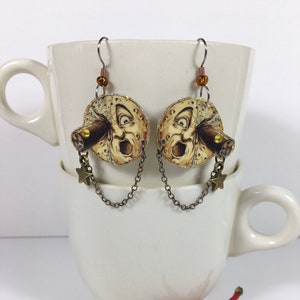 May include: A pair of gold-toned earrings featuring a round image of a man's face with a large mouth and a telescope. The earrings are attached to a chain with a star charm.